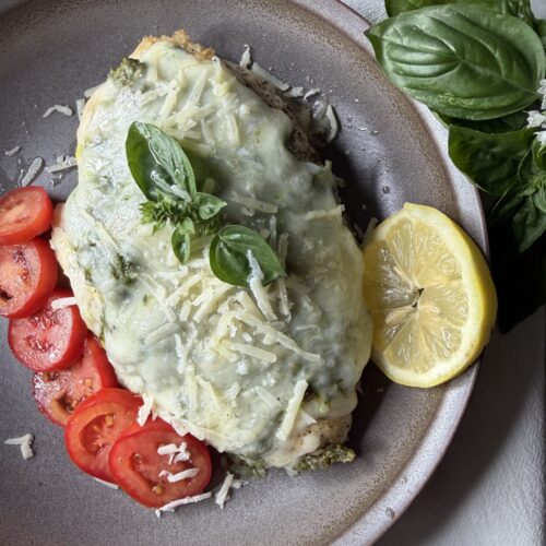 Chicken breast coated with fresh garden pesto and topped with savory cheeses on a plate served with sliced tomatoes.