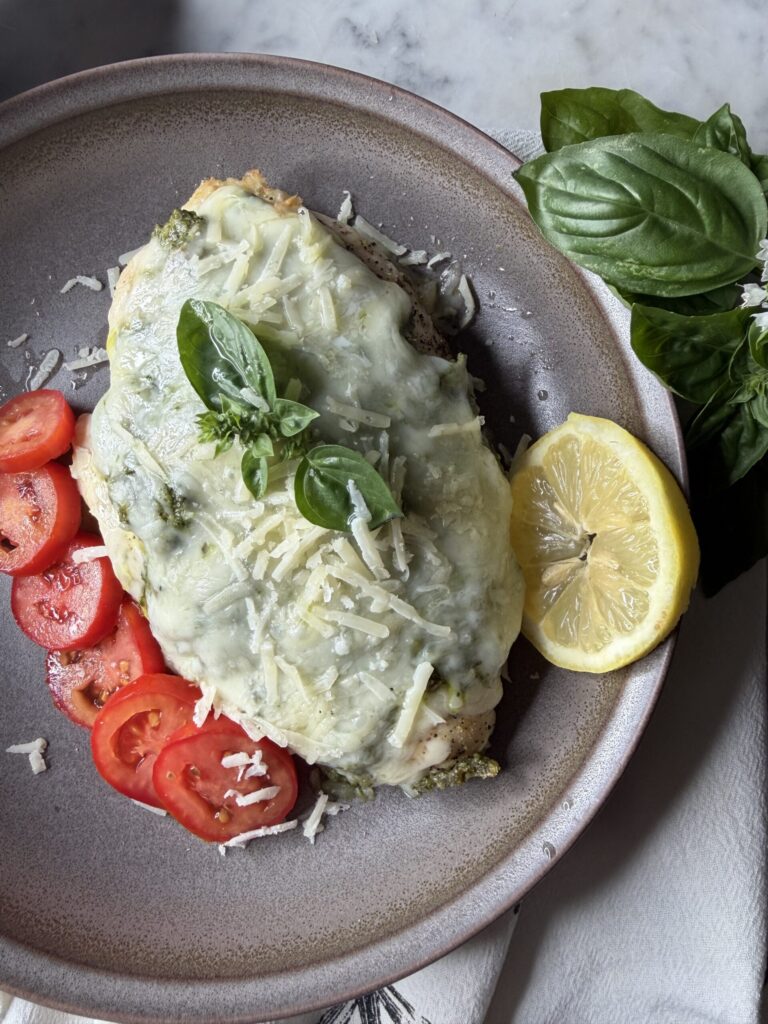 Chicken breast coated with fresh garden pesto and topped with savory cheeses on a plate served with sliced tomatoes.