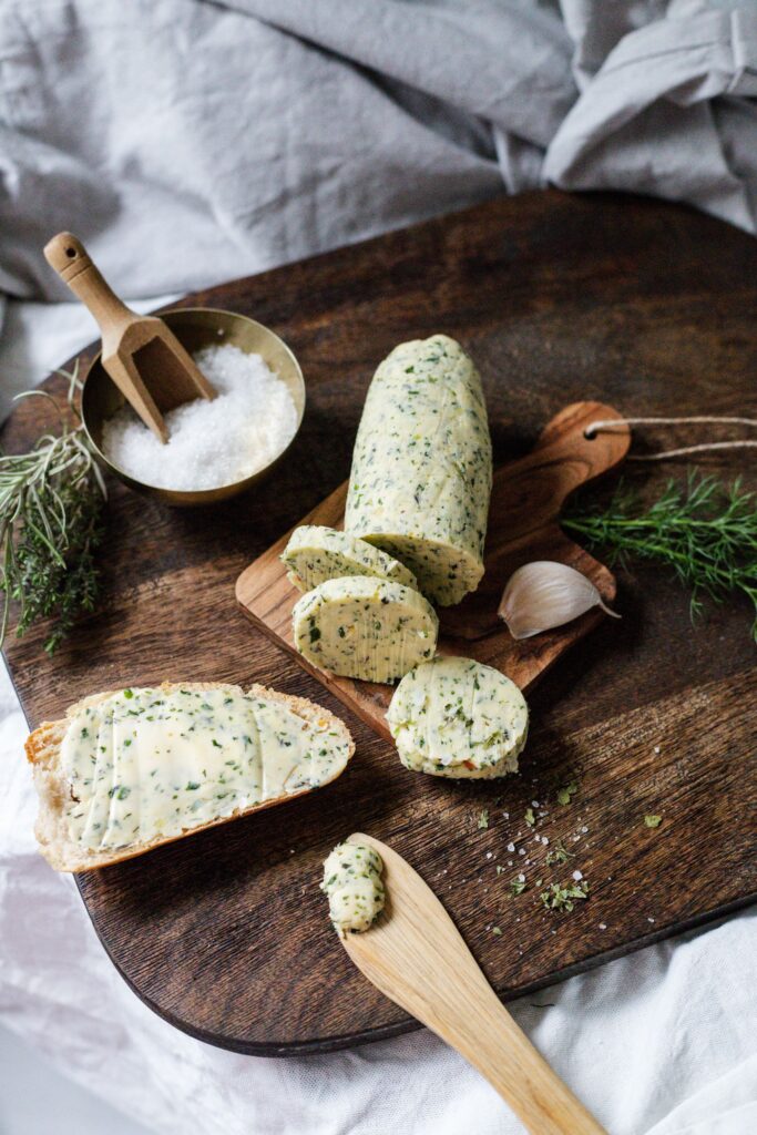 A rustic wooden board featuring herb-infused butter spread on bread, with garlic and salt.