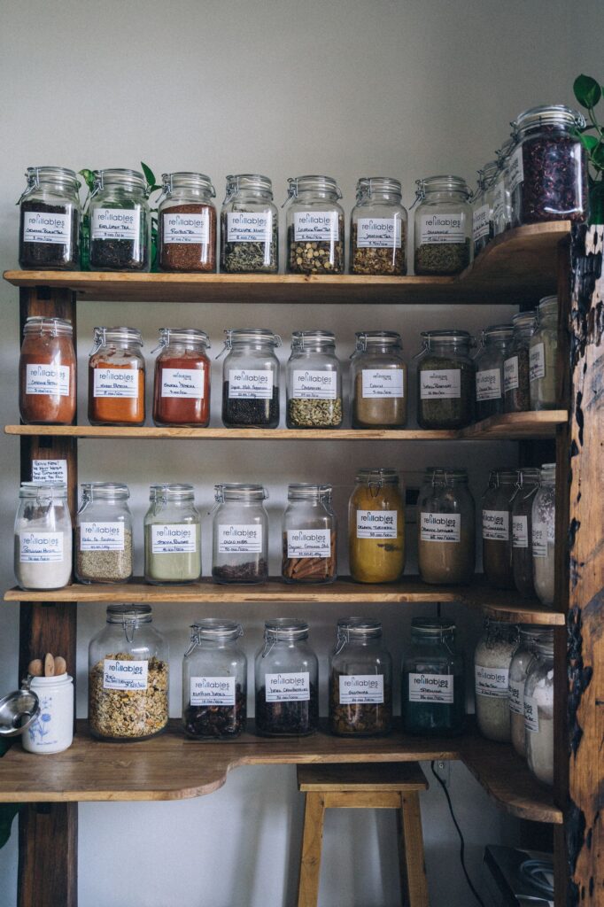 A wooden shelf displaying assorted spices in clear refillable glass jars, encouraging eco-friendly storage.