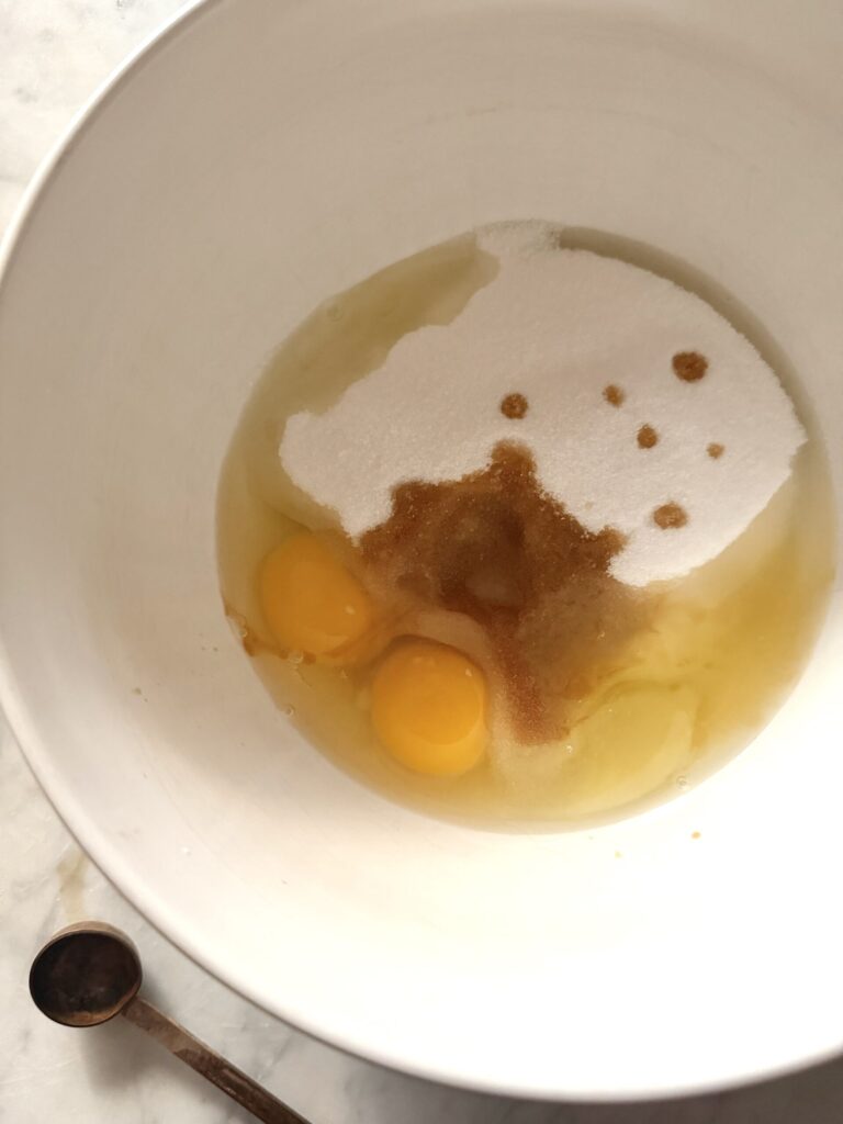 Eggs, vanilla, and granulated sugar are added to a white mixing bowl.