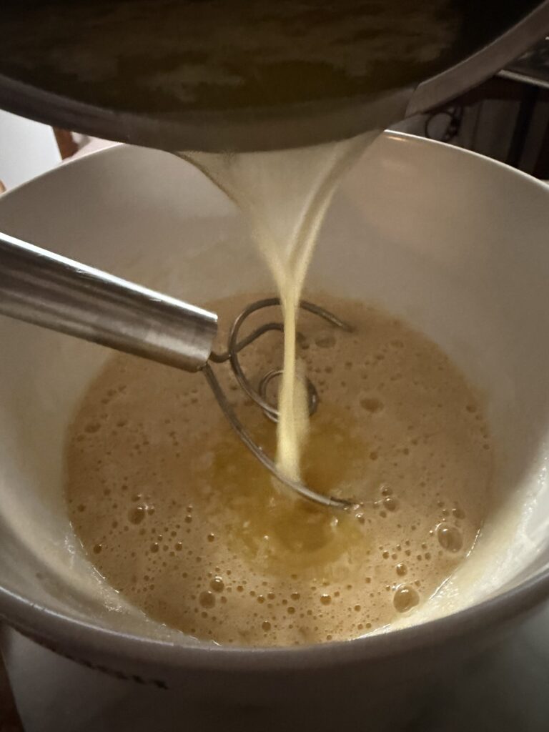 A warm mixture of melted butter and sugar is added to a bowl of sugar, vanilla, and eggs to make brownie batter.