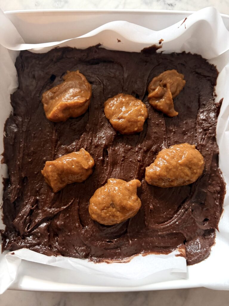 Spoonfuls of a pumpkin mixture are added to the top of a pan of brownie batter ready to swirl.