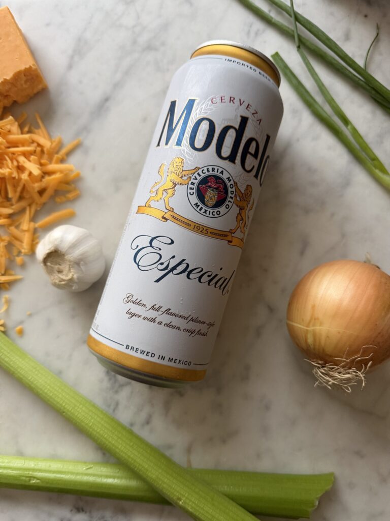 Beer can, onion, scallions, celery, garlic cloves, and shredded cheddar cheese on a marble surface