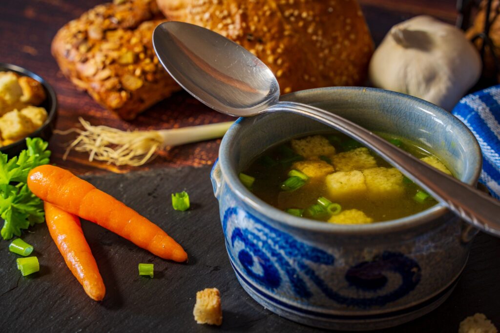 A cozy scene of vegetable broth with croutons, fresh carrots, bread, and herbs, perfect for a comforting meal.